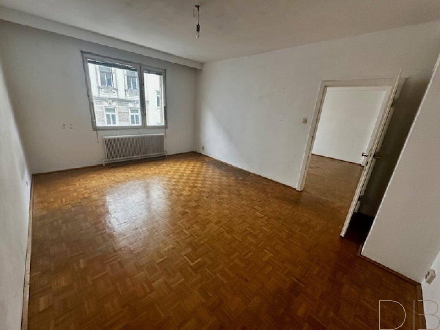 Geräumiges Zimmer mit Parkettboden und Fensterblick auf die umliegenden Gebäude.