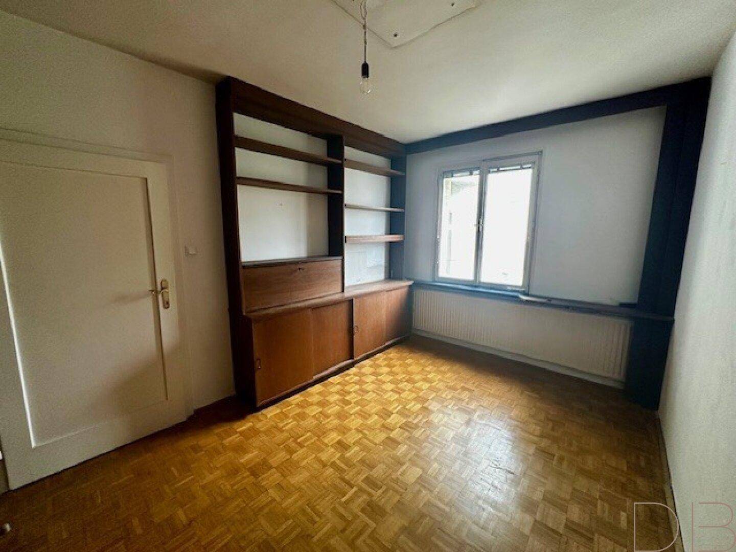 Zimmer mit Einbauregal, Parkettboden und Fenster, das viel Tageslicht hereinlässt.