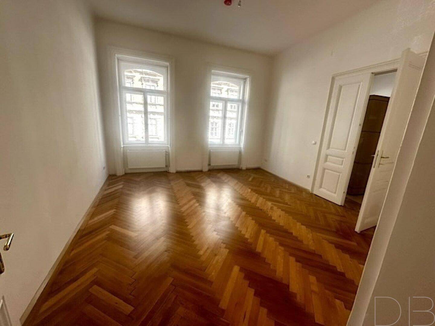 Geräumiges Zimmer mit klassischem Fischgrätparkett und zwei großen Fenstern.