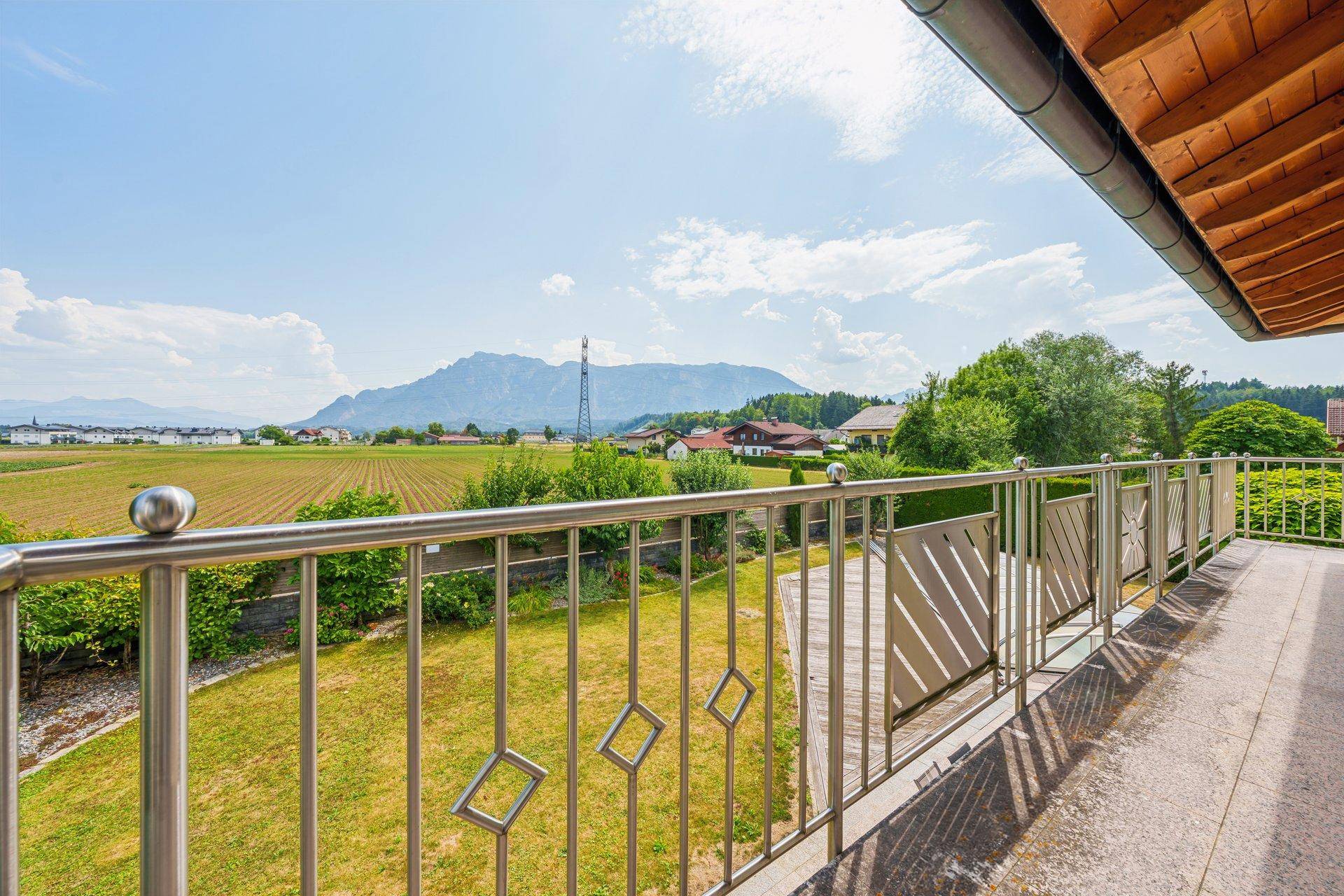 Balkon mit Metallgeländer und weitem Blick über Felder und Berge unter blauem Himmel.