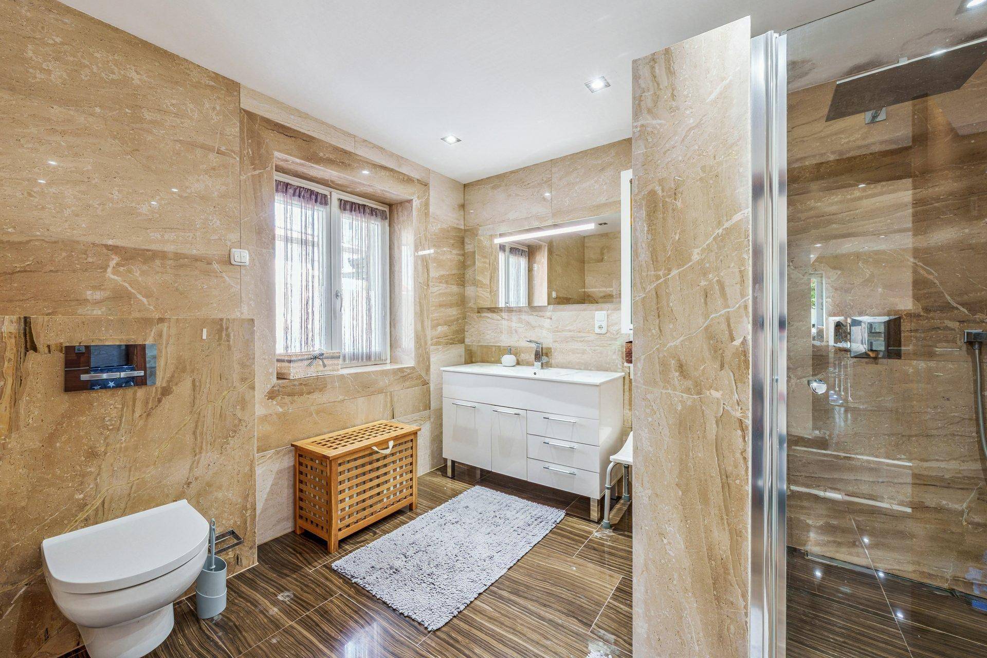 Modernes Badezimmer mit beigen Marmorfliesen, begehbarer Dusche und einem Fenster für Tageslicht.