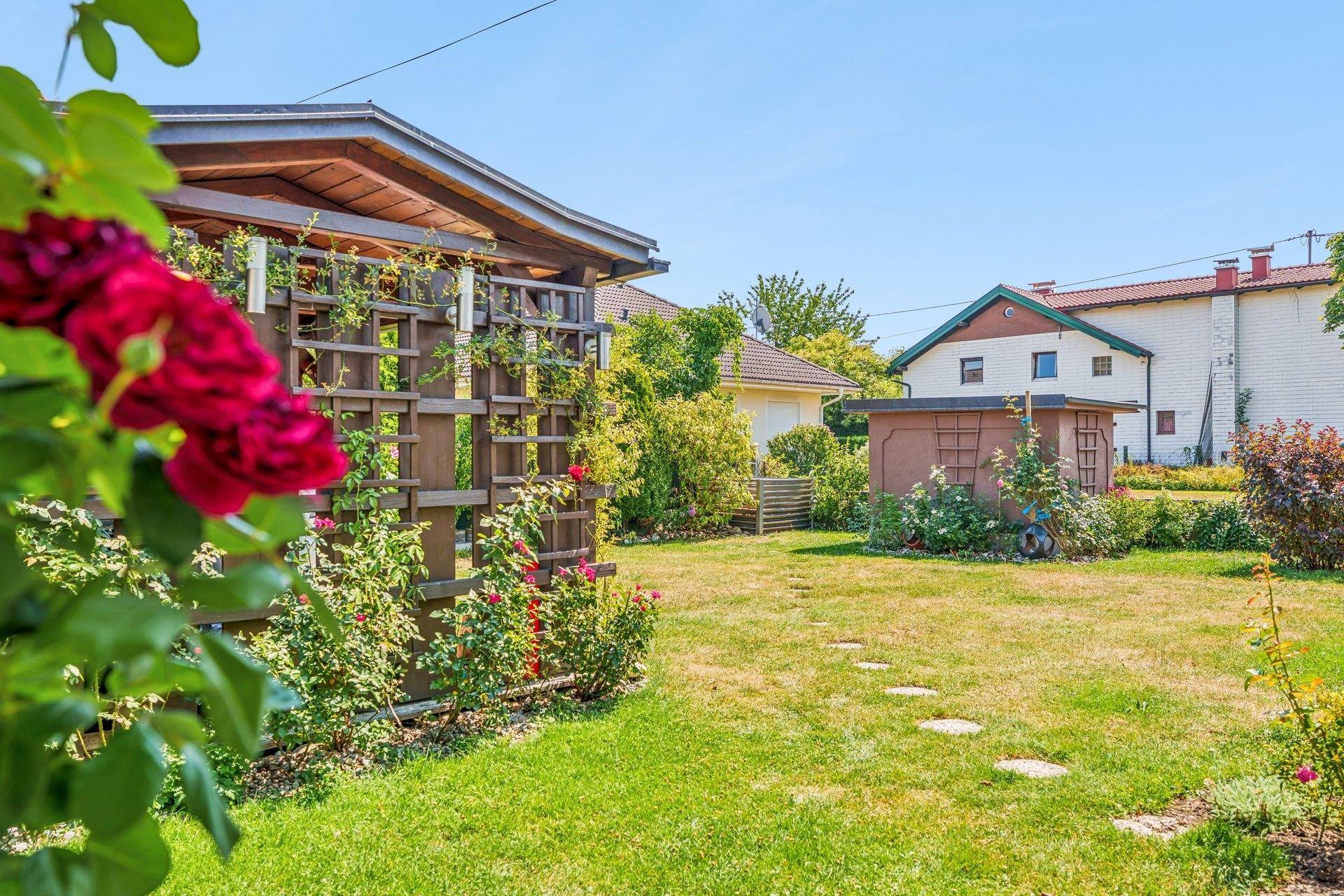 Gepflegter Garten mit Rasenfläche, Rosenranken an einer Holzpergola und einem kleinen Gartenhaus.