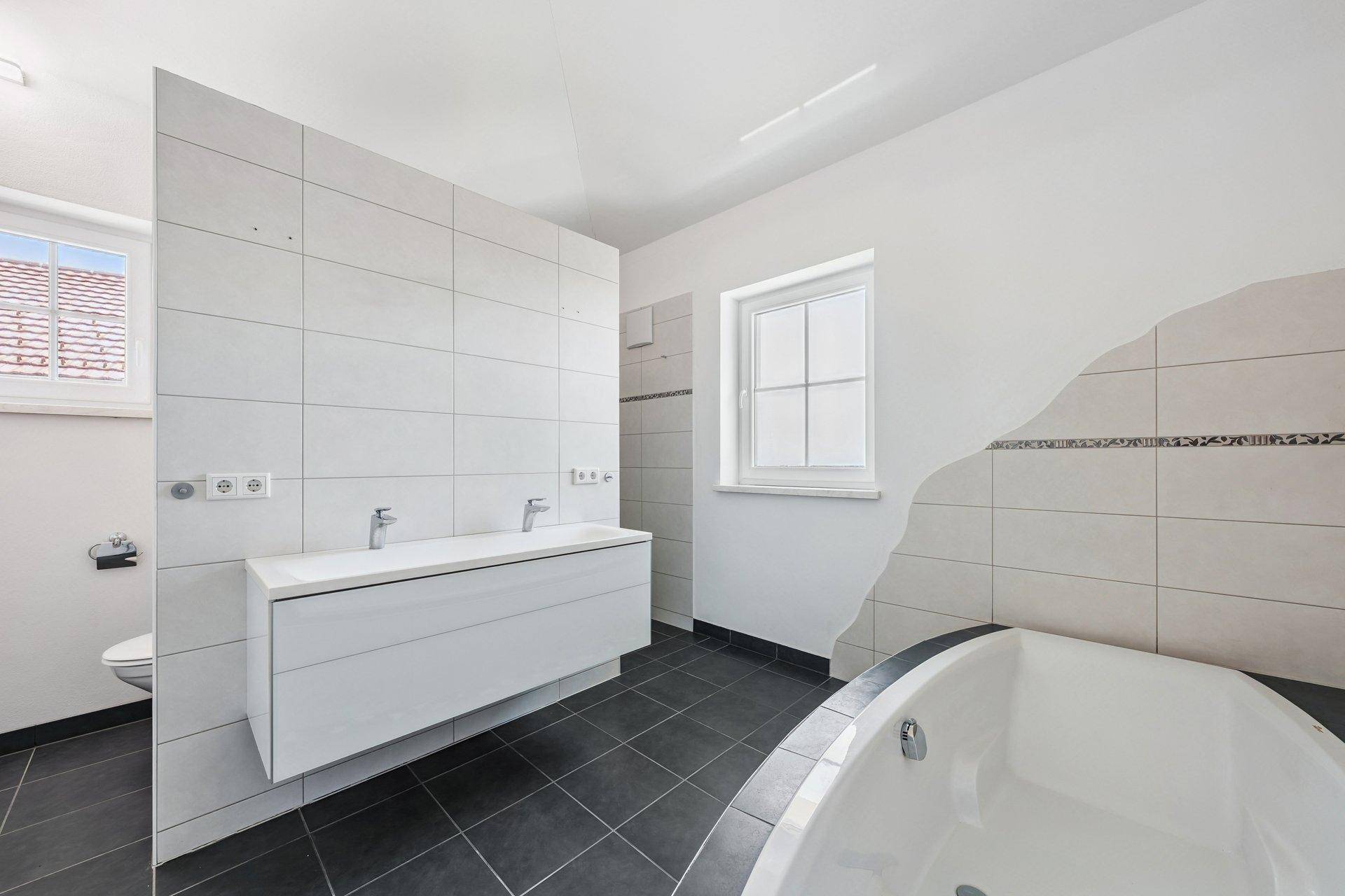 Modernes Badezimmer mit Doppelwaschbecken, Badewanne und dunklen Fliesen.