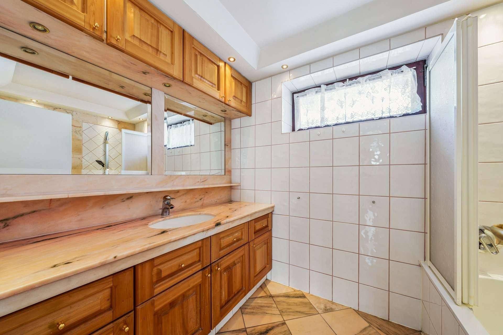 Badezimmer mit Marmorwaschtisch, Holzschränken, Fliesenboden und einem Fenster.