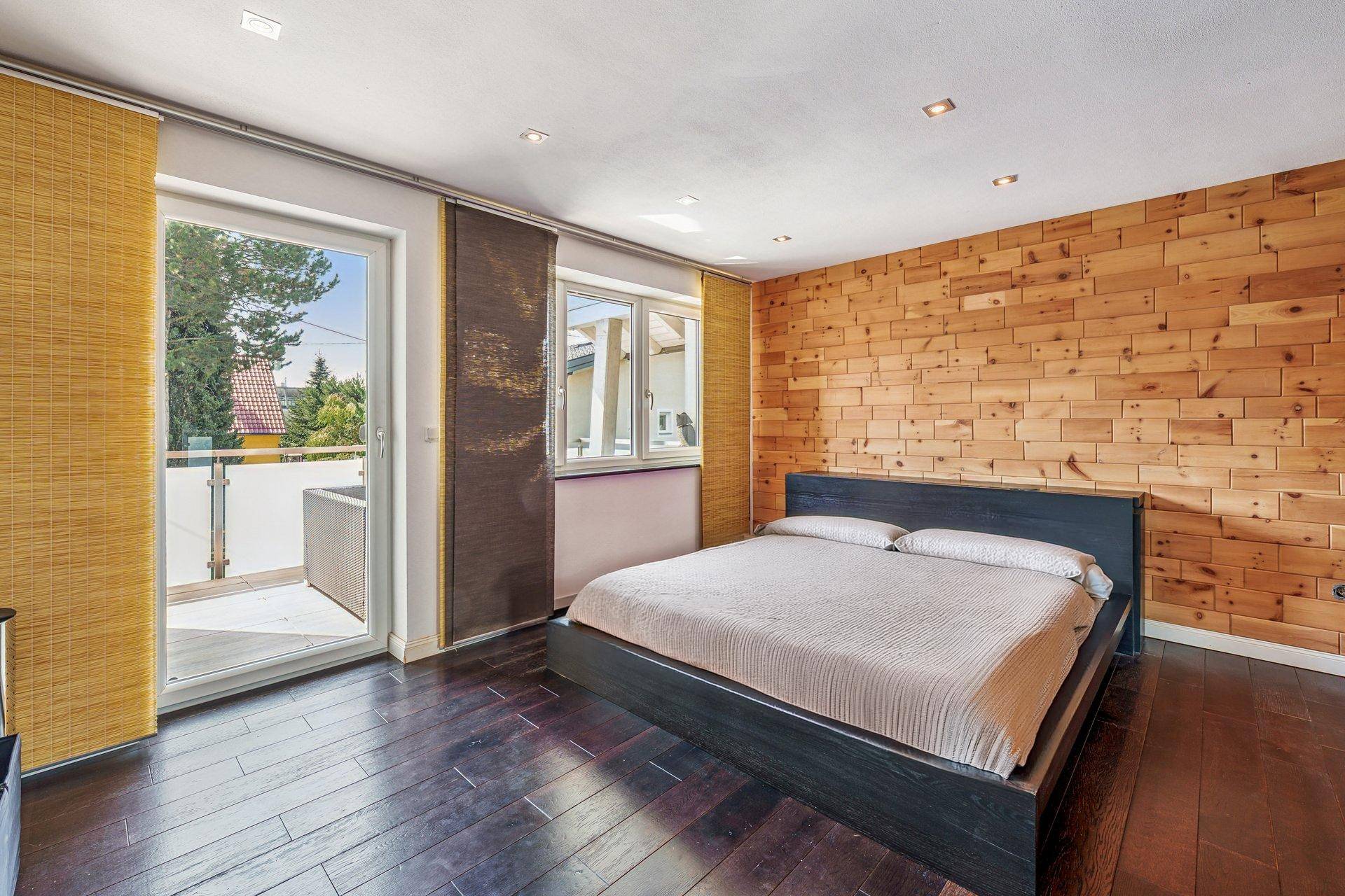 Schlafzimmer mit dunklem Holzboden, Holzwandakzent und Zugang zu einem Balkon mit Blick ins Grüne.