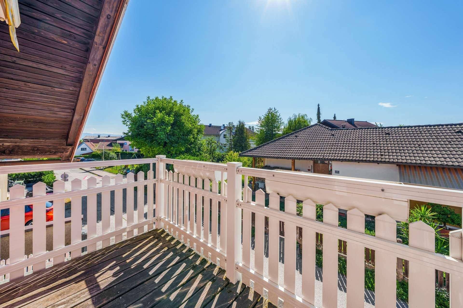 Sonniger Balkon mit weißem Holzgeländer, Holzboden und Blick auf die grüne Umgebung.