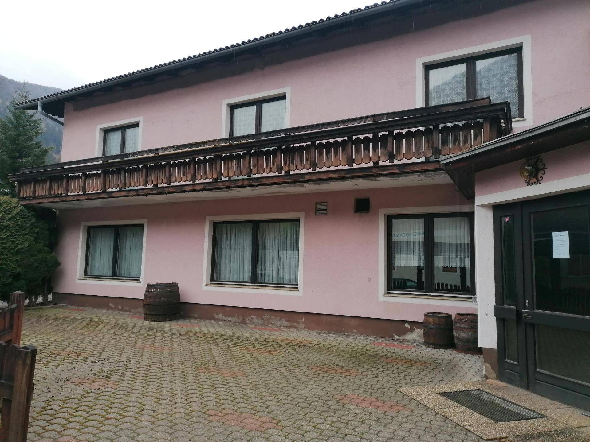 Rosa gestrichenes Gebäude mit traditionellem Holzbalkon und gepflastertem Vorplatz, umgeben von Bäumen.