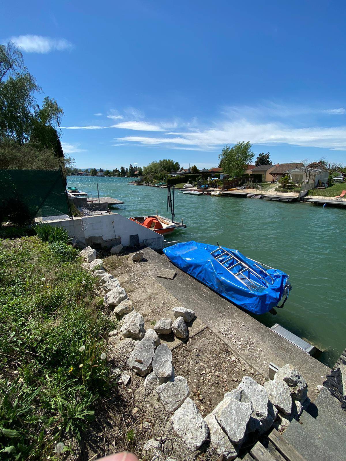 Blick vom Ufer auf den Kanal mit einem blauen Boot und felsigen Elementen im Vordergrund.