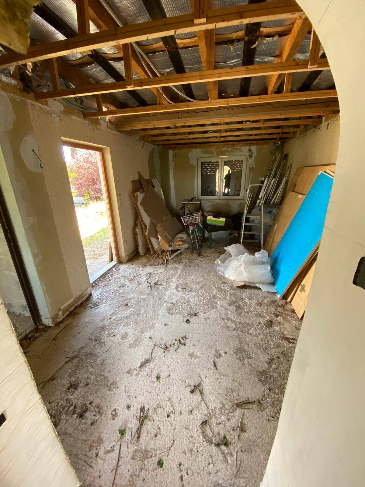 Innenraum im Rohbau mit freiliegenden Holzbalken, Betonboden und einem Fenster.