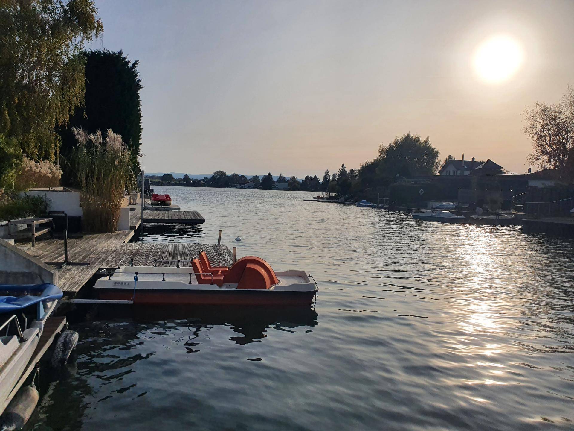 Stimmungsvoller Sonnenuntergang über dem Kanal mit einem Tretboot am Steg und Häusern am Horizont.