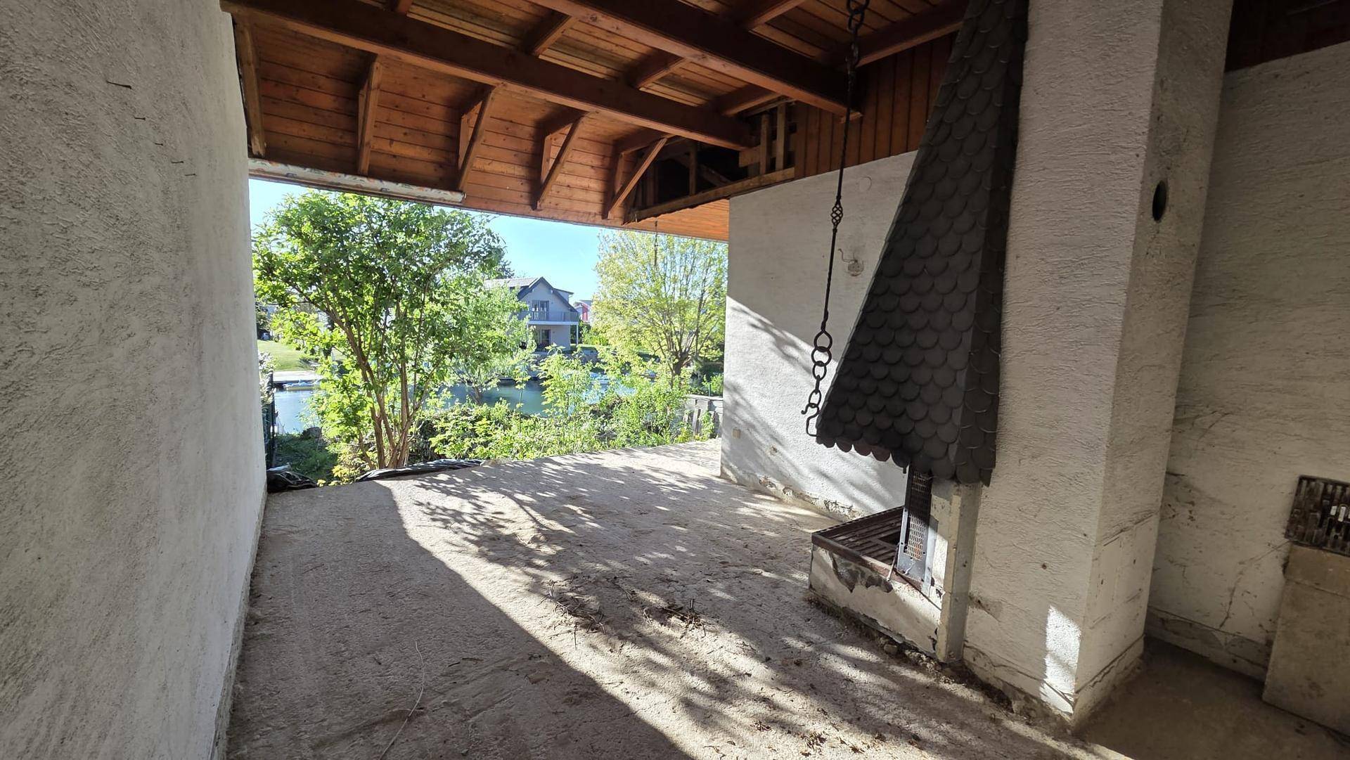 Überdachter Außenbereich mit gemauertem Grillkamin, Betonboden und Holzdecke, Blick zum Kanal.