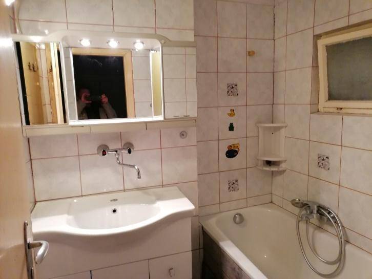 Badezimmer mit weißen Fliesen, Waschbecken, Spiegelschrank und Badewanne mit Duschfunktion sowie einem Fenster.
