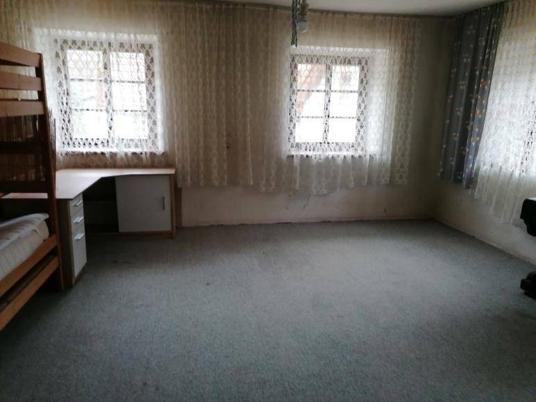 Schlafzimmer mit grauem Teppichboden, zwei Fenstern mit Spitzenvorhängen und einem Schreibtischbereich.