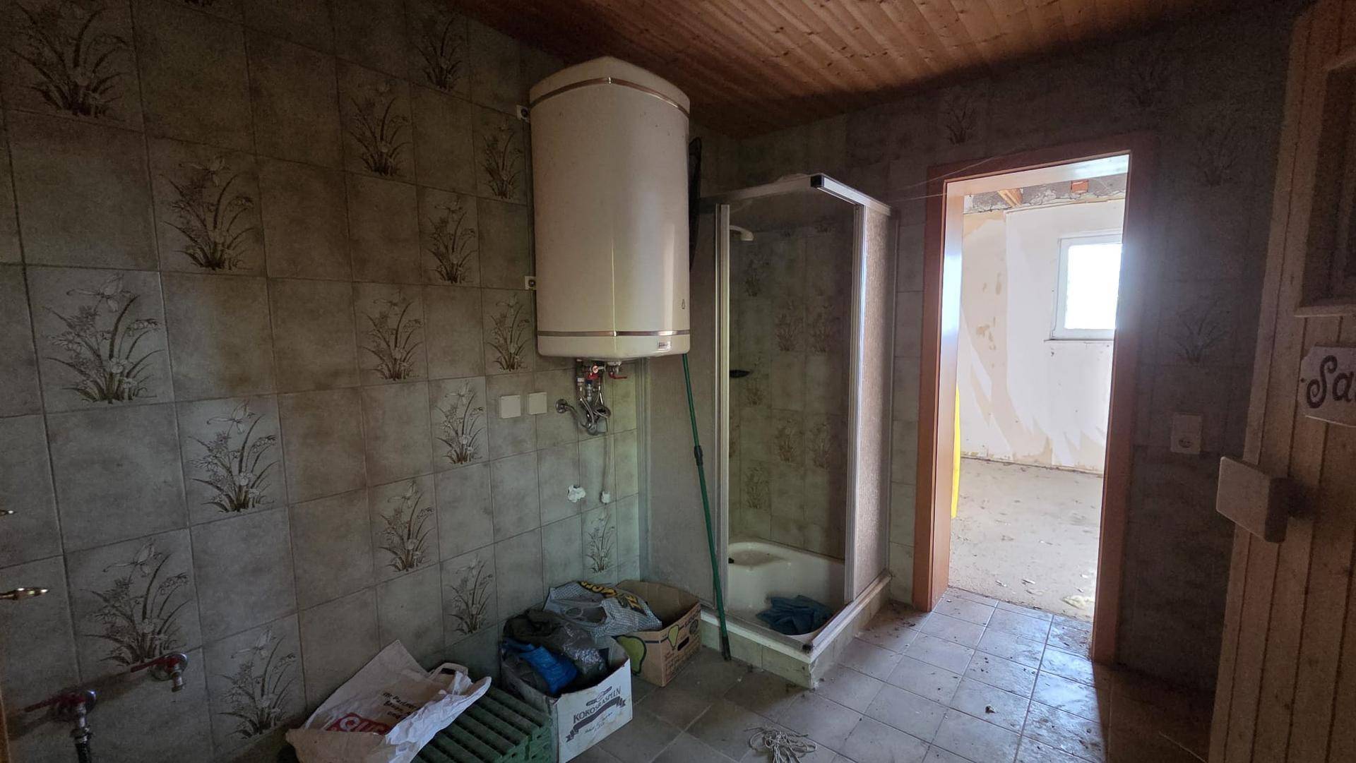 Badezimmer im Renovierungszustand mit alten Fliesen, Warmwasserboiler und Duschkabine.