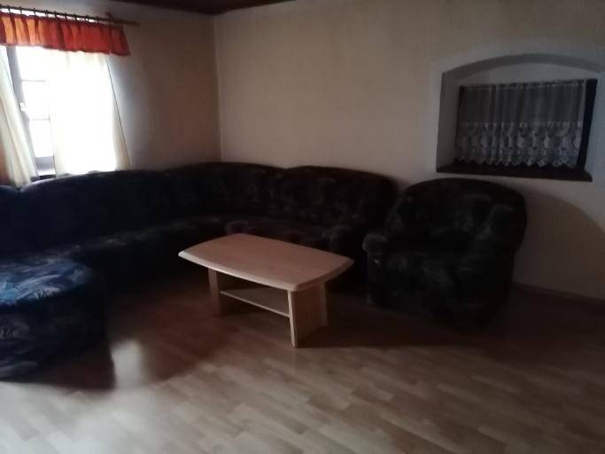 Wohnzimmer mit dunklem Ecksofa, Couchtisch, Laminatboden und zwei Fenstern, die Tageslicht hereinlassen.
