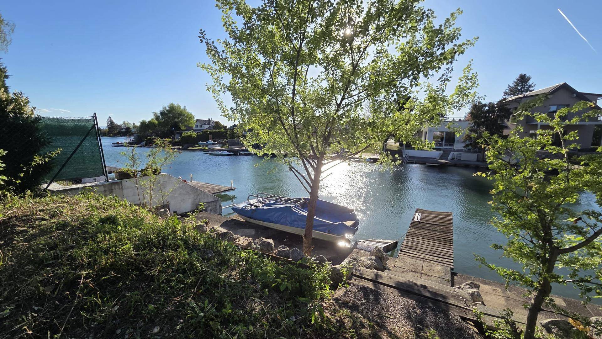 Grüne Uferlandschaft mit einem Baum, einem Boot am Wasser und Häusern am gegenüberliegenden Ufer.