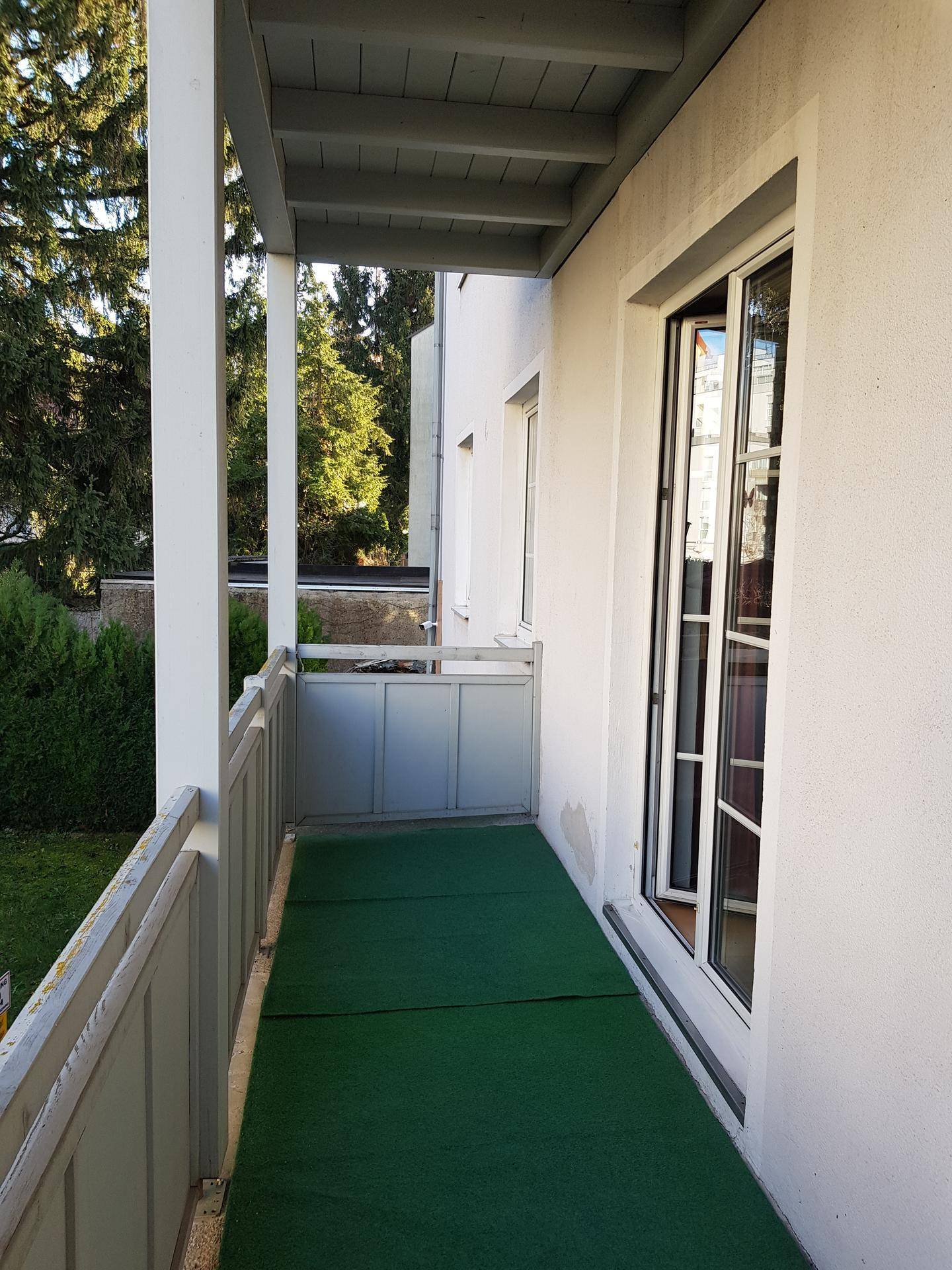 Schmaler Balkon mit grünem Kunstrasen, Geländer und Blick ins Grüne mit Bäumen.