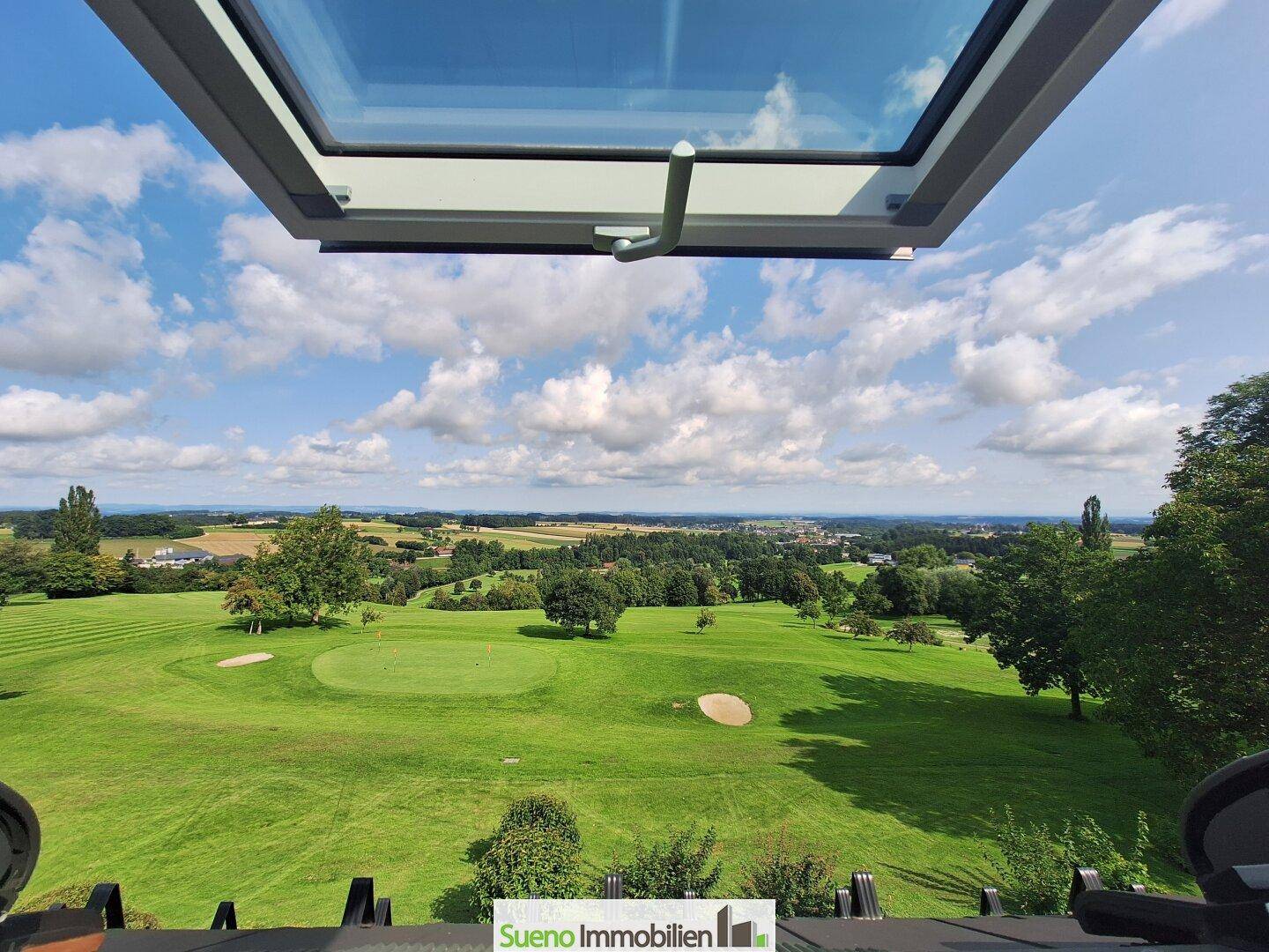 Weitläufiger Blick aus einem Dachfenster auf eine grüne Golfplatzlandschaft unter blauem Himmel.