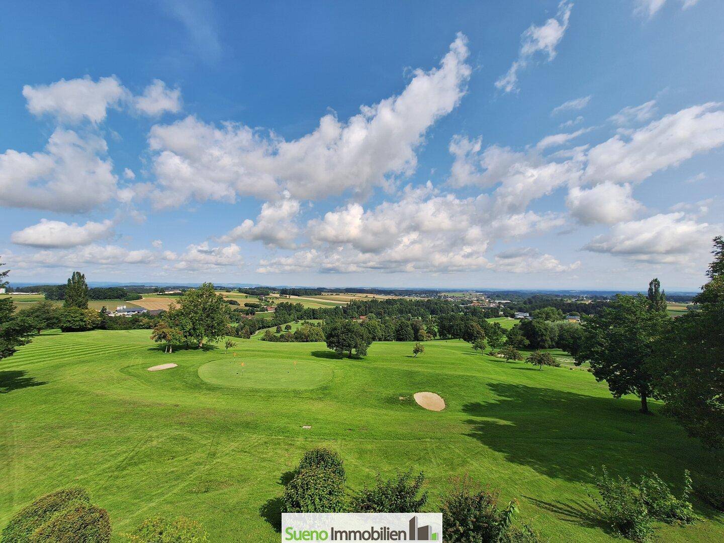 Panoramablick auf eine weitläufige Golfplatzlandschaft mit Bäumen und blauem Himmel.
