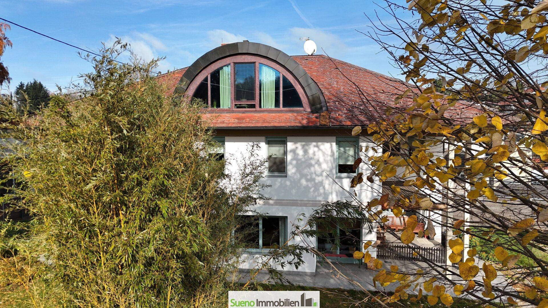 Vorderansicht des Hauses mit markantem Dachfenster und Terrasse, umgeben von herbstlichem Grün.