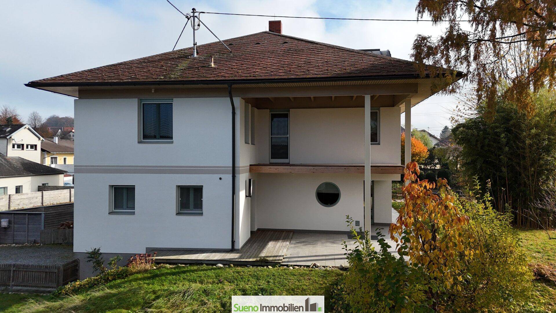 Seitliche Ansicht des Hauses mit Balkon und runden Fenstern, umgeben von herbstlicher Vegetation.