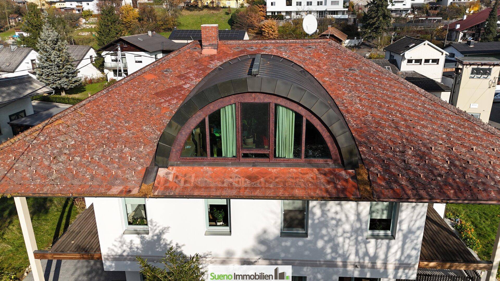 Detailansicht des Daches mit markantem Rundbogenfenster und Ziegeldeckung, umgeben von Bäumen.