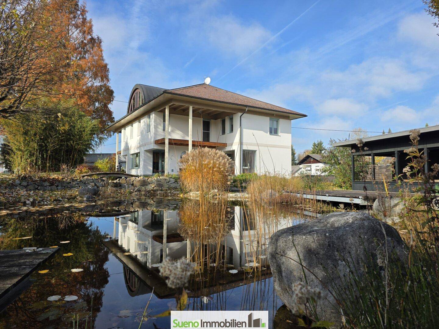 Großzügiges Einfamilienhaus mit weitläufigem Garten und idyllischem Schwimmteich unter blauem Himmel.