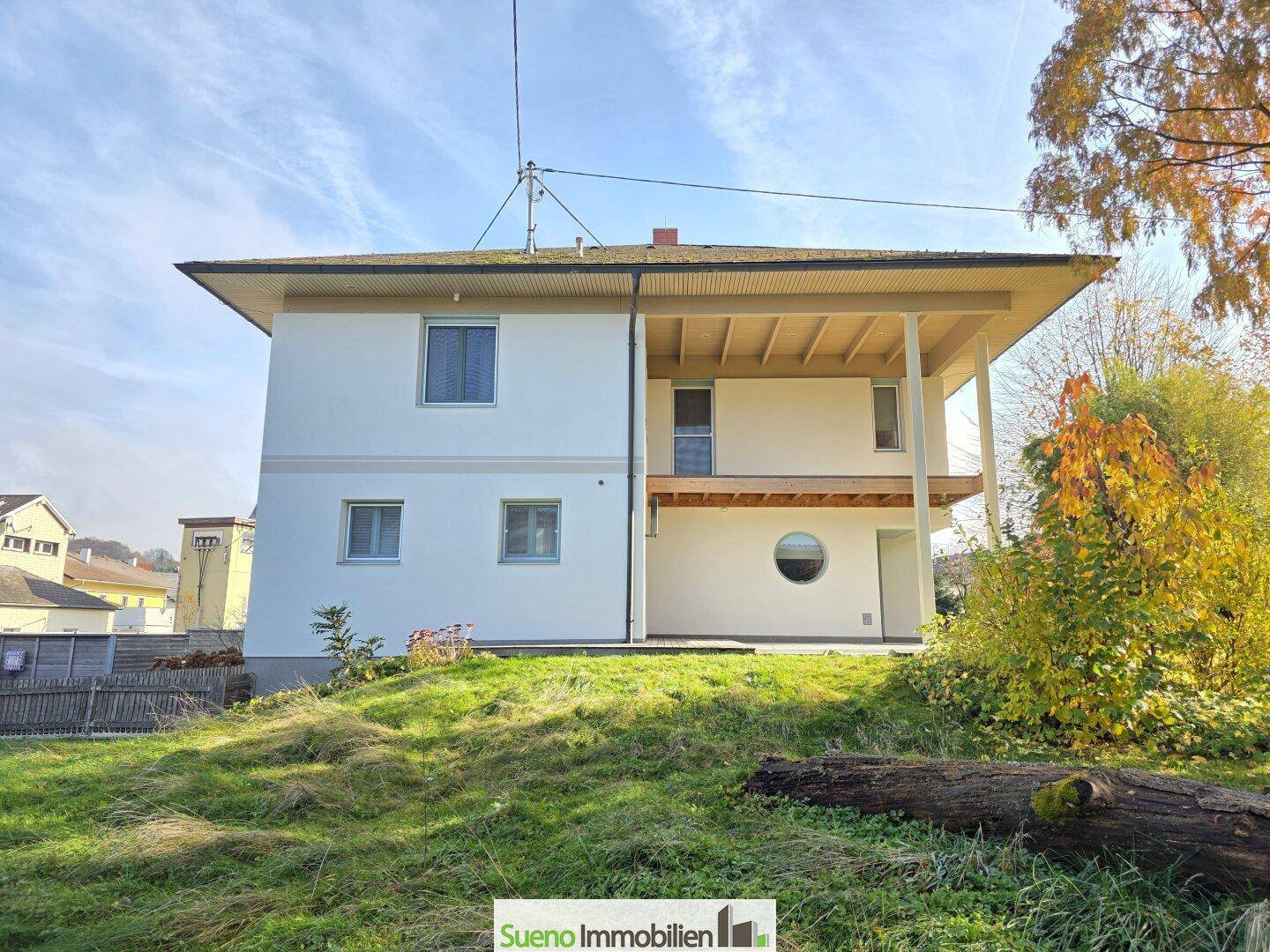 Seitliche Ansicht des Hauses mit Balkon und runden Fenstern, umgeben von herbstlicher Vegetation.