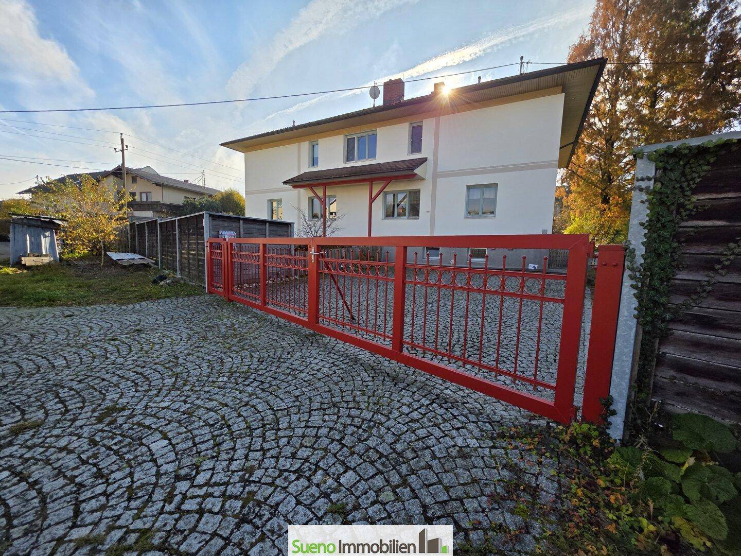 Rotes Metalltor und gepflasterte Zufahrt zum Anwesen, umgeben von herbstlicher Natur.