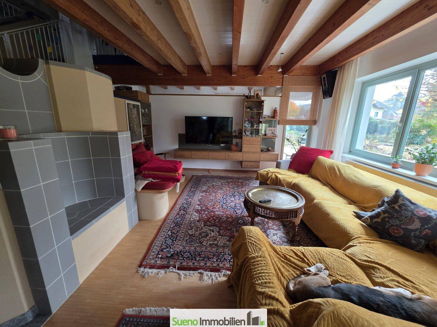Gemütliches Wohnzimmer mit Holzbalkendecke, Kachelofen und bequemen Sofas in warmen Farben.
