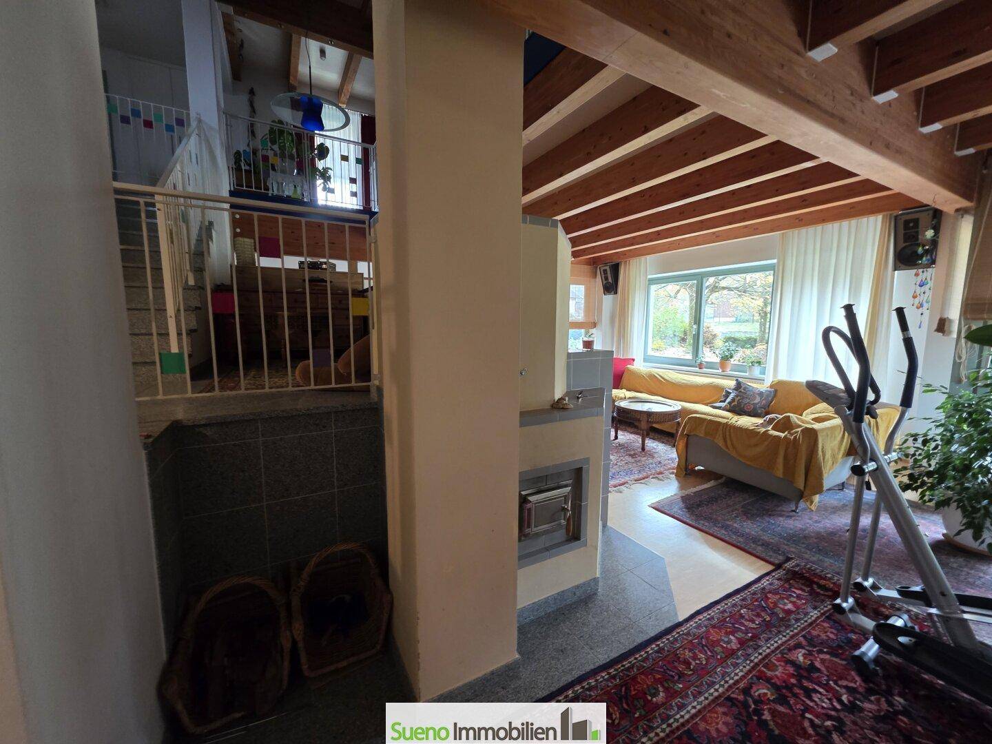 Eingangsbereich mit Treppe und Blick in das helle Wohnzimmer mit Kachelofen und Holzbalken.