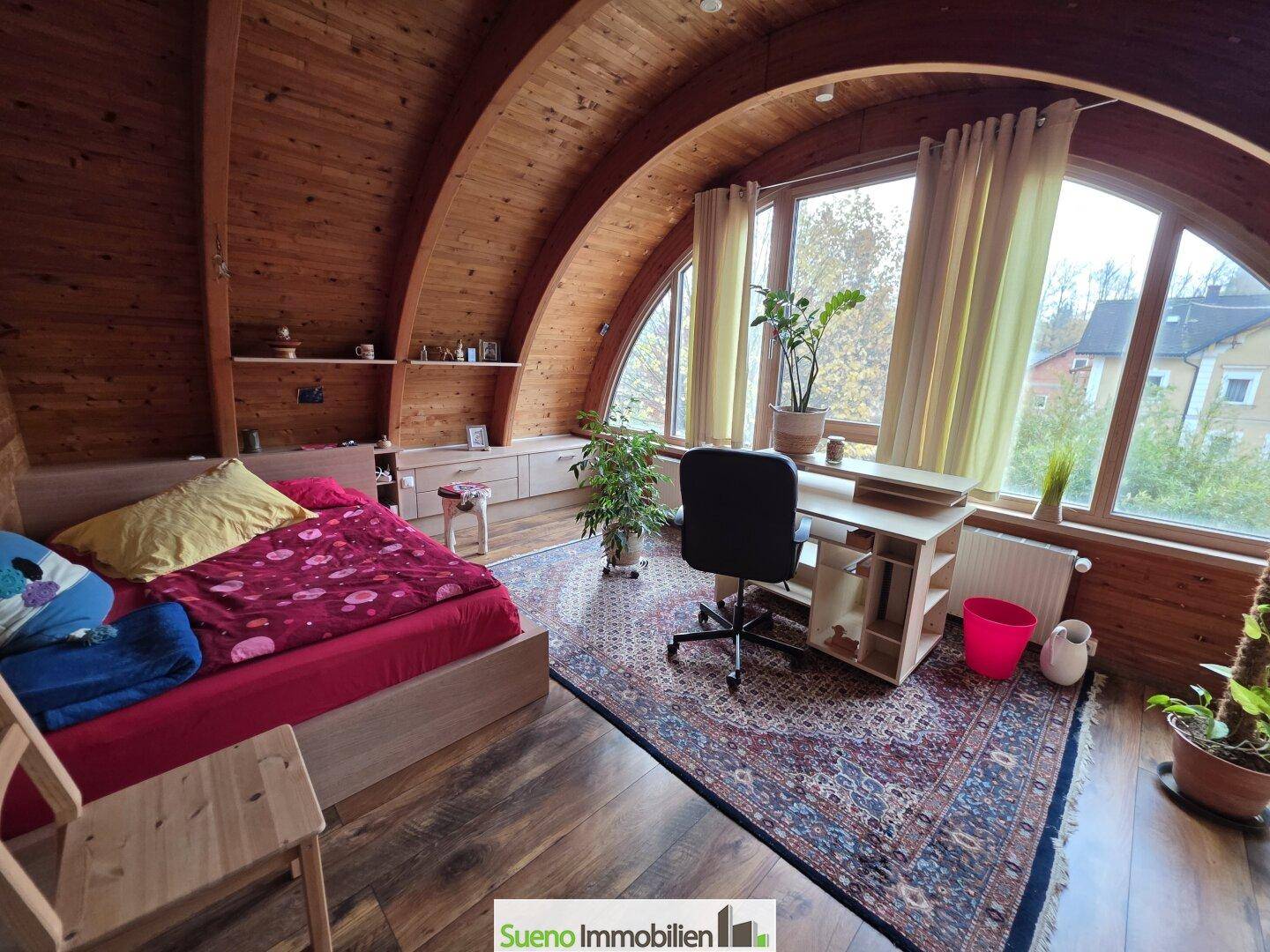 Schlafzimmer im Dachgeschoss mit Holzwänden, großen Fenstern und Arbeitsbereich.