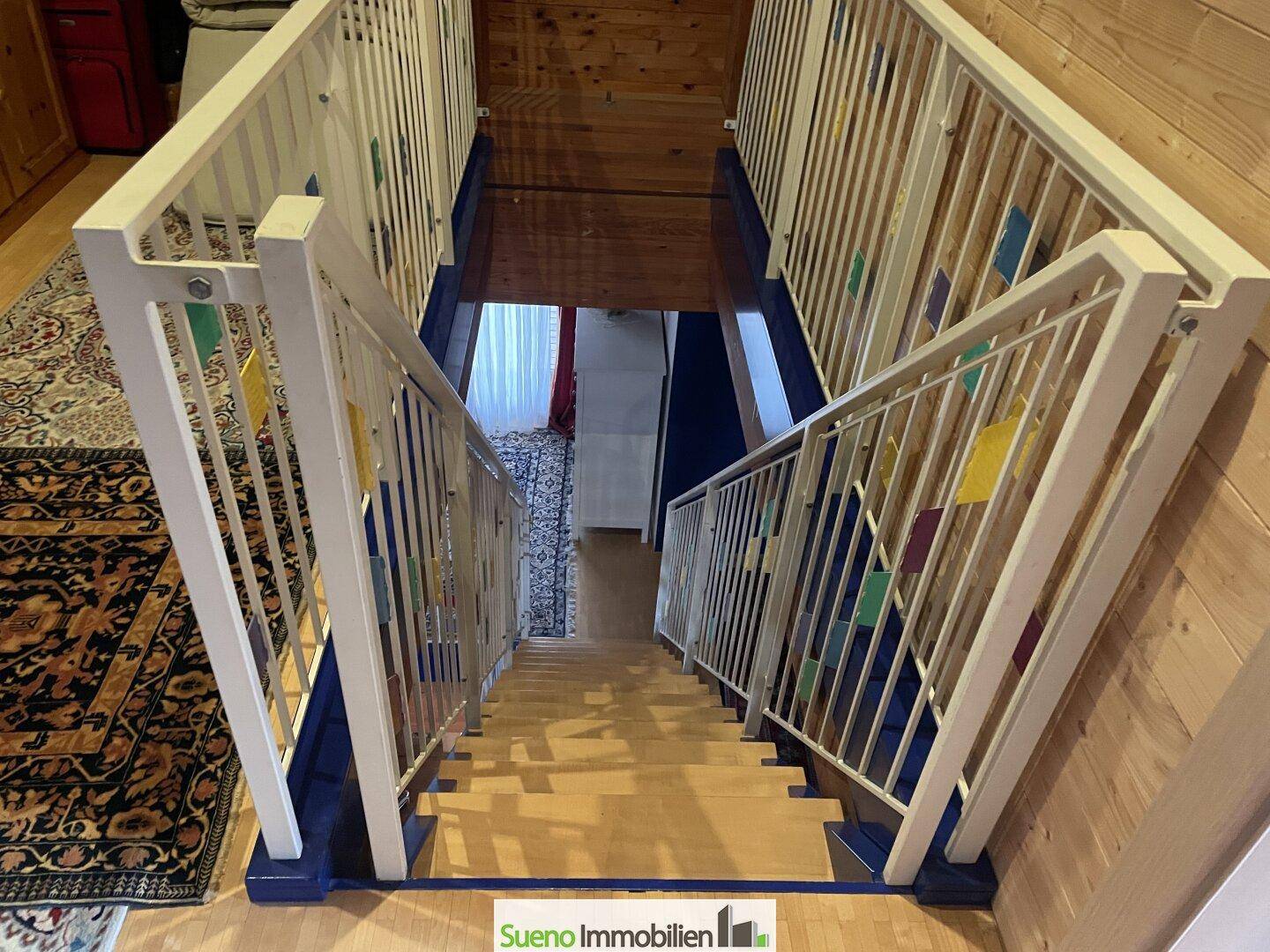 Treppenhaus mit Holzstufen, weißem Geländer und bunten Dekorationselementen.