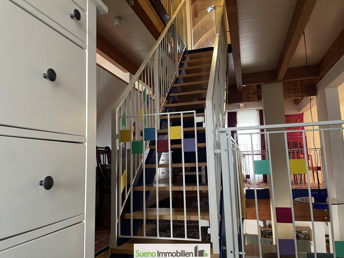 Treppenhaus mit Holzstufen, weißem Geländer und bunten Akzenten, führt zu oberen Ebenen.