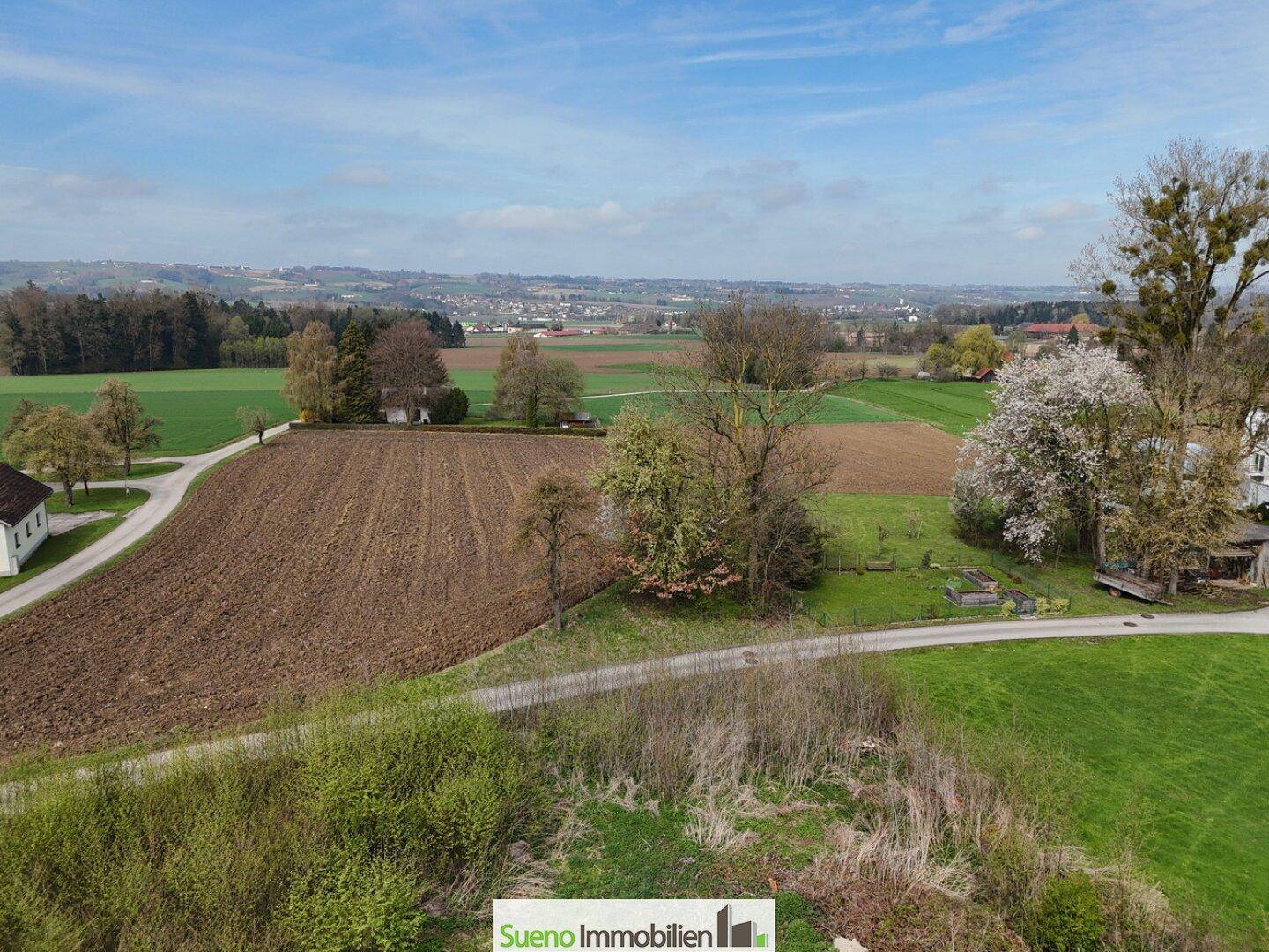 Weitläufige ländliche Landschaft mit Feldern, Bäumen und einem Dorf in der Ferne unter blauem Himmel.