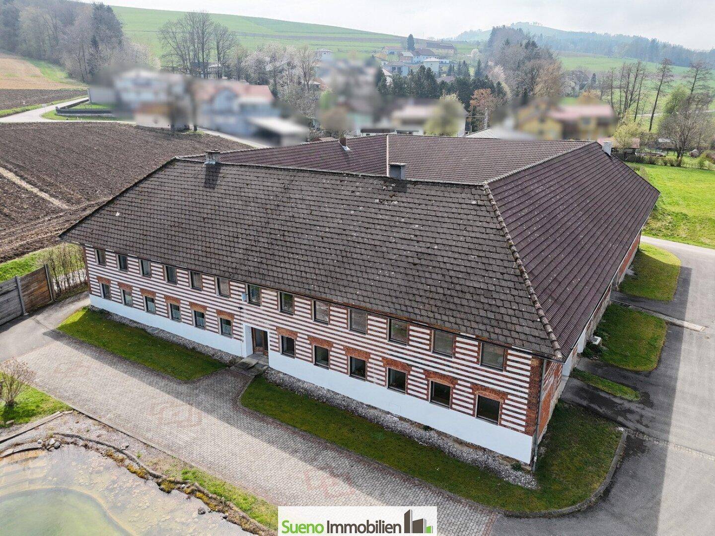 Luftaufnahme des großen Bauernhauses mit umliegenden Feldern und ländlicher Bebauung im Hintergrund.