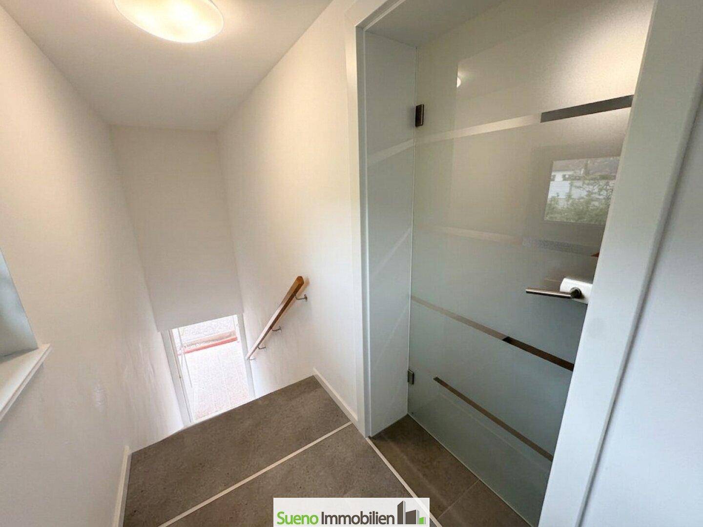 Heller Treppenaufgang mit Holzgeländer und Glastür, die zu einem weiteren Raum führt.