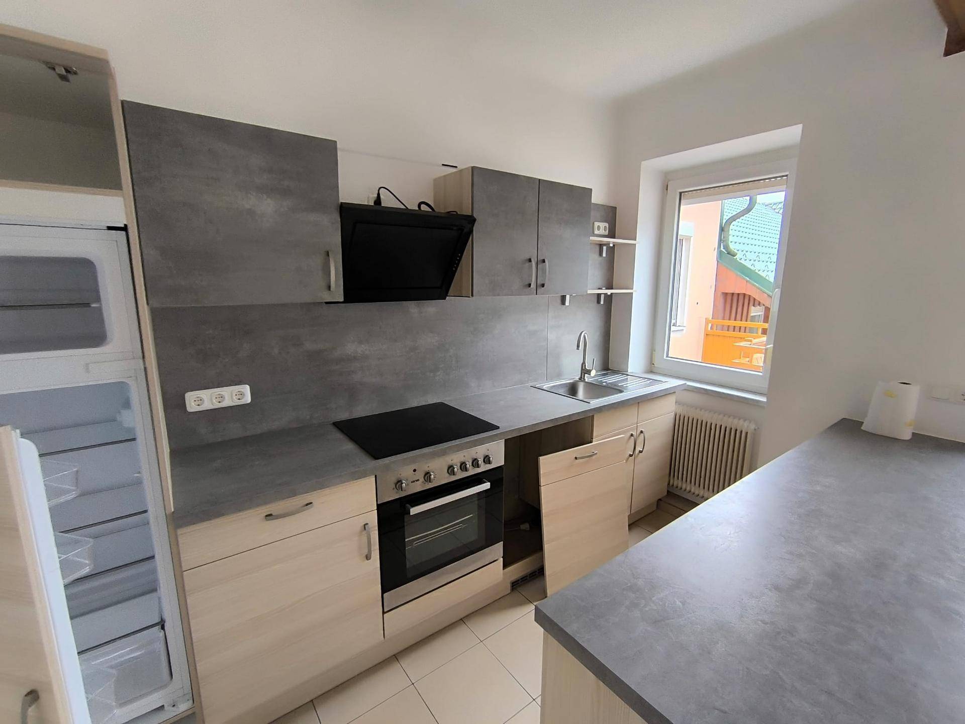 Moderne Küche mit hellen Holzfronten, grauen Arbeitsplatten und integrierten Geräten, inklusive Kühlschrank und Kochfeld.