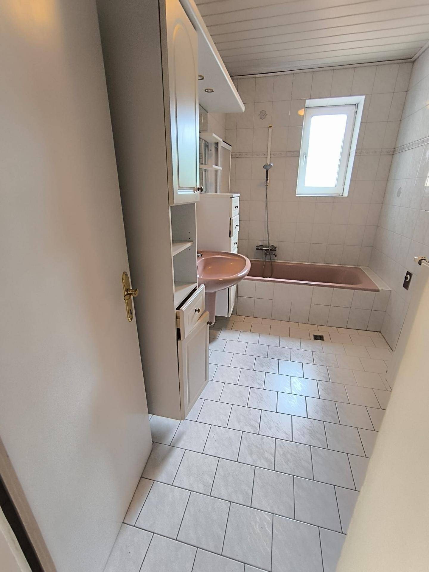 Helles Badezimmer mit rosa Badewanne, weißen Fliesen und einem Fenster für Tageslicht.