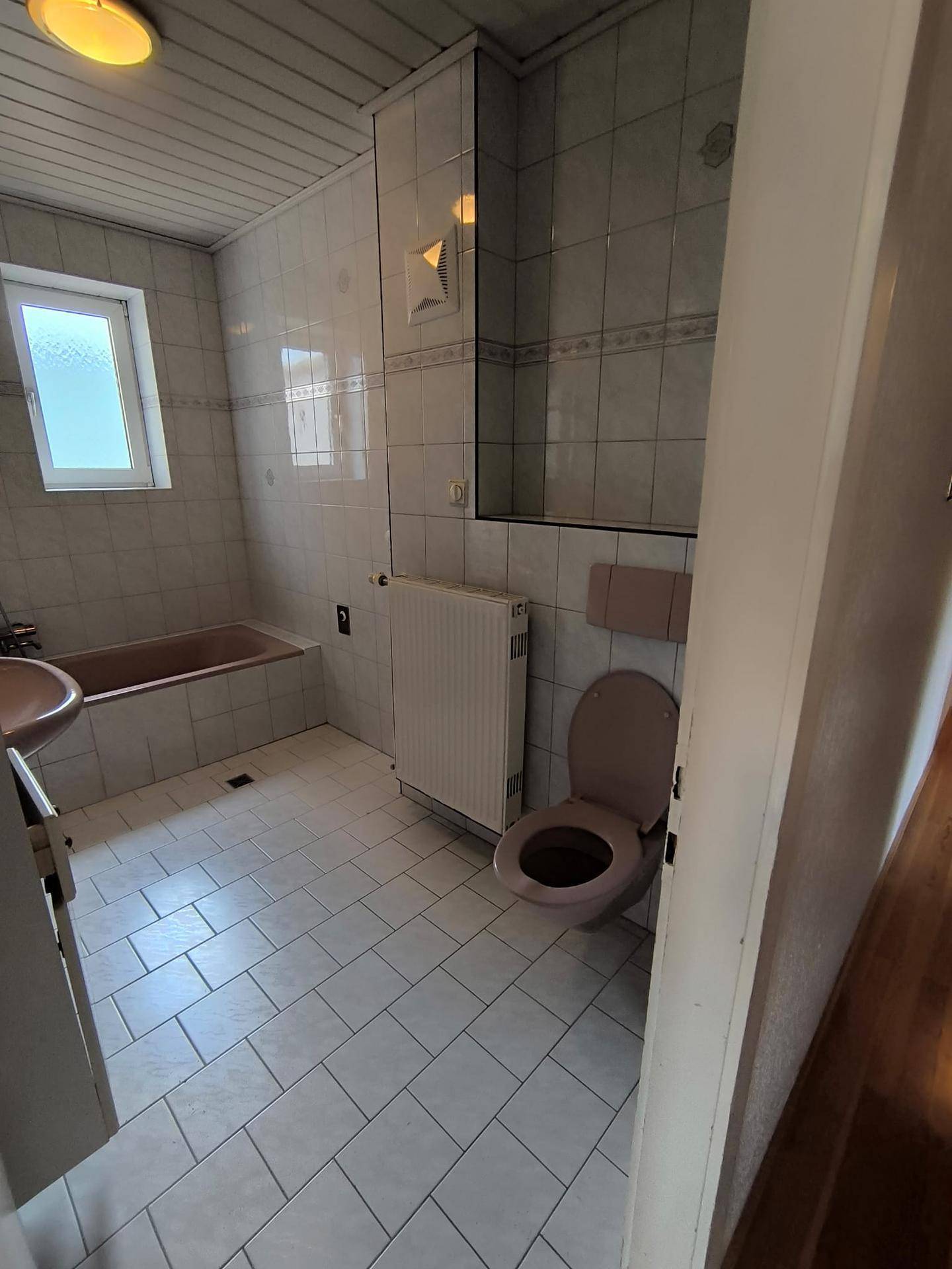 Badezimmer mit WC, rosa Badewanne und Fenster, ausgestattet mit weißen Fliesen und Holzdecke.