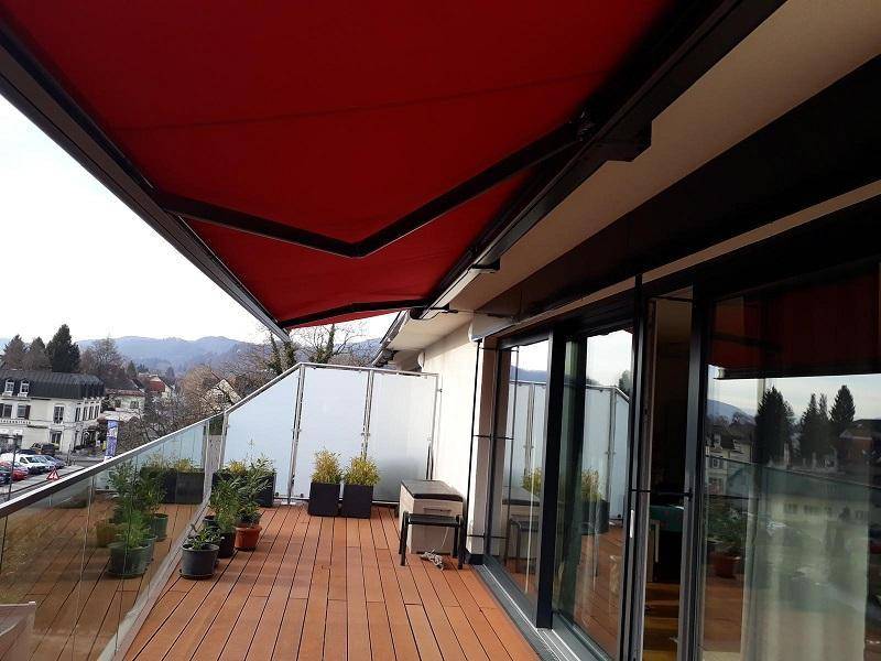 Sonniger Terrassenbereich mit ausgefahrener roter Markise, Holzbelag und Panoramablick auf die Stadt und Berge.