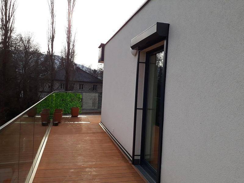 Lange Terrasse mit Holzbelag und Glasgeländer, die sich entlang der modernen Gebäudefassade erstreckt.