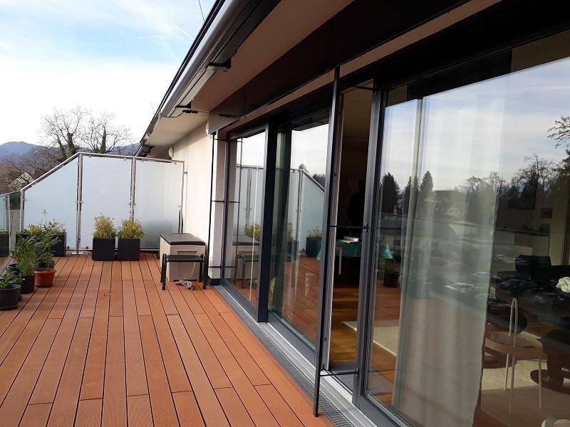 Weitläufige Terrasse mit Holzplankenboden und raumhohen Glasfronten, die viel Tageslicht in den Innenraum lassen.