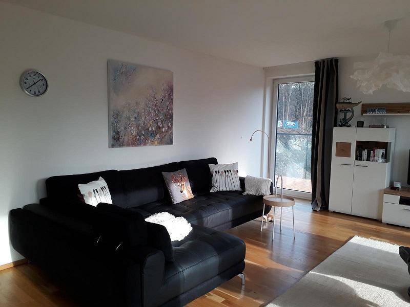 Helles Wohnzimmer mit großem schwarzem Ecksofa, Holzparkett und Zugang zu einem sonnigen Außenbereich.