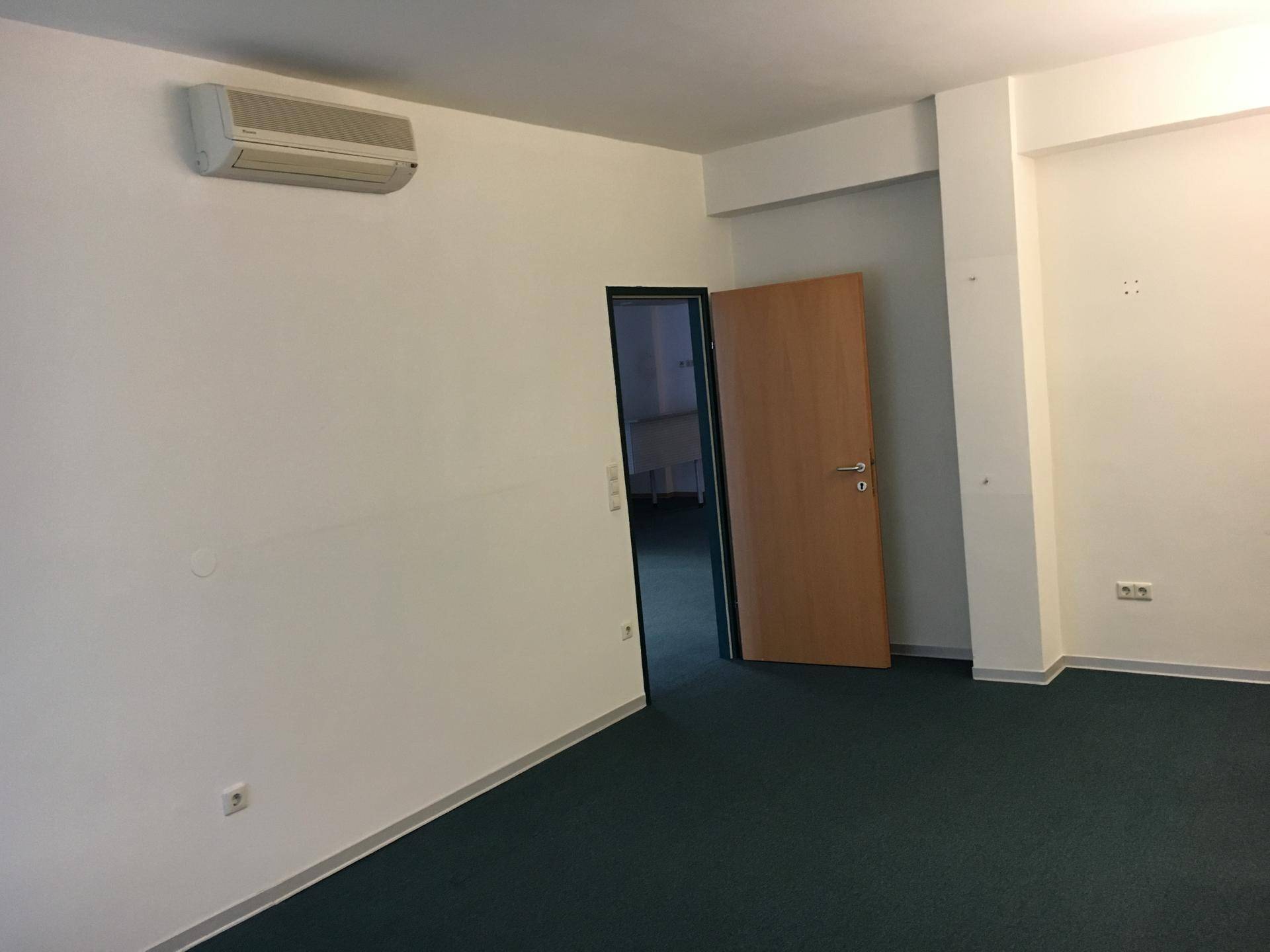 Büroraum mit Teppichboden, Klimaanlage und einer offenen Holztür zu einem weiteren Bereich.