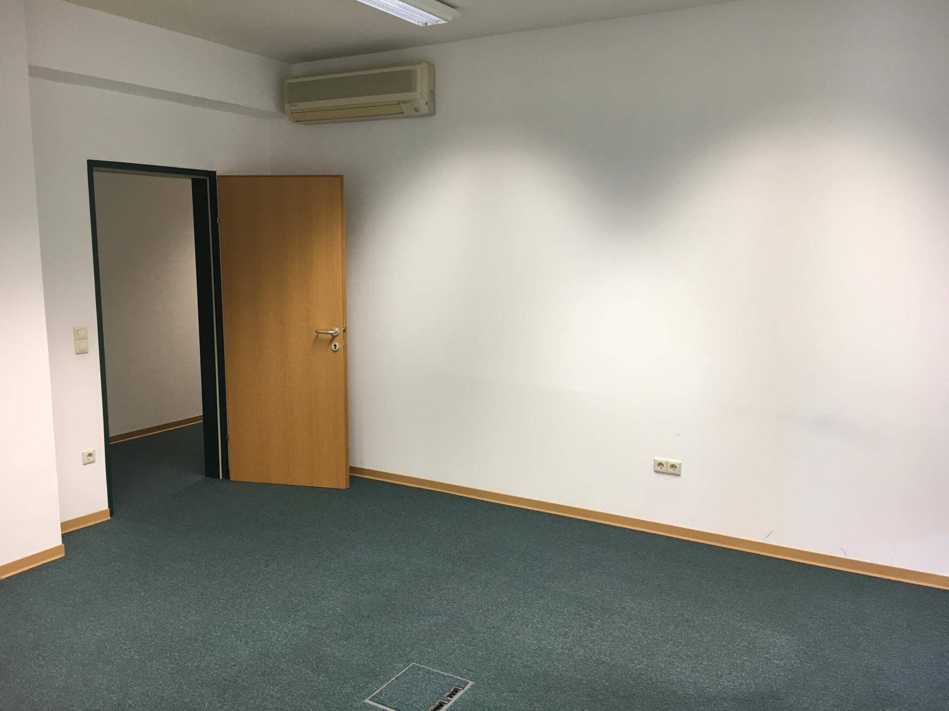 Büroraum mit Klimaanlage, offener Holztür und einem Bodentank im Teppichboden.