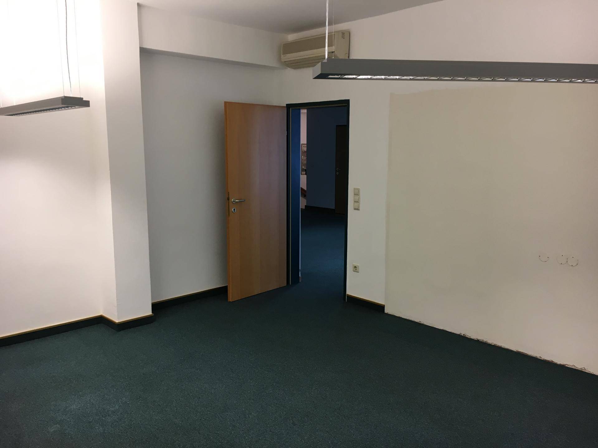 Büroraum mit Teppichboden, Klimaanlage und einer offenen Holztür zu einem weiteren Bereich.