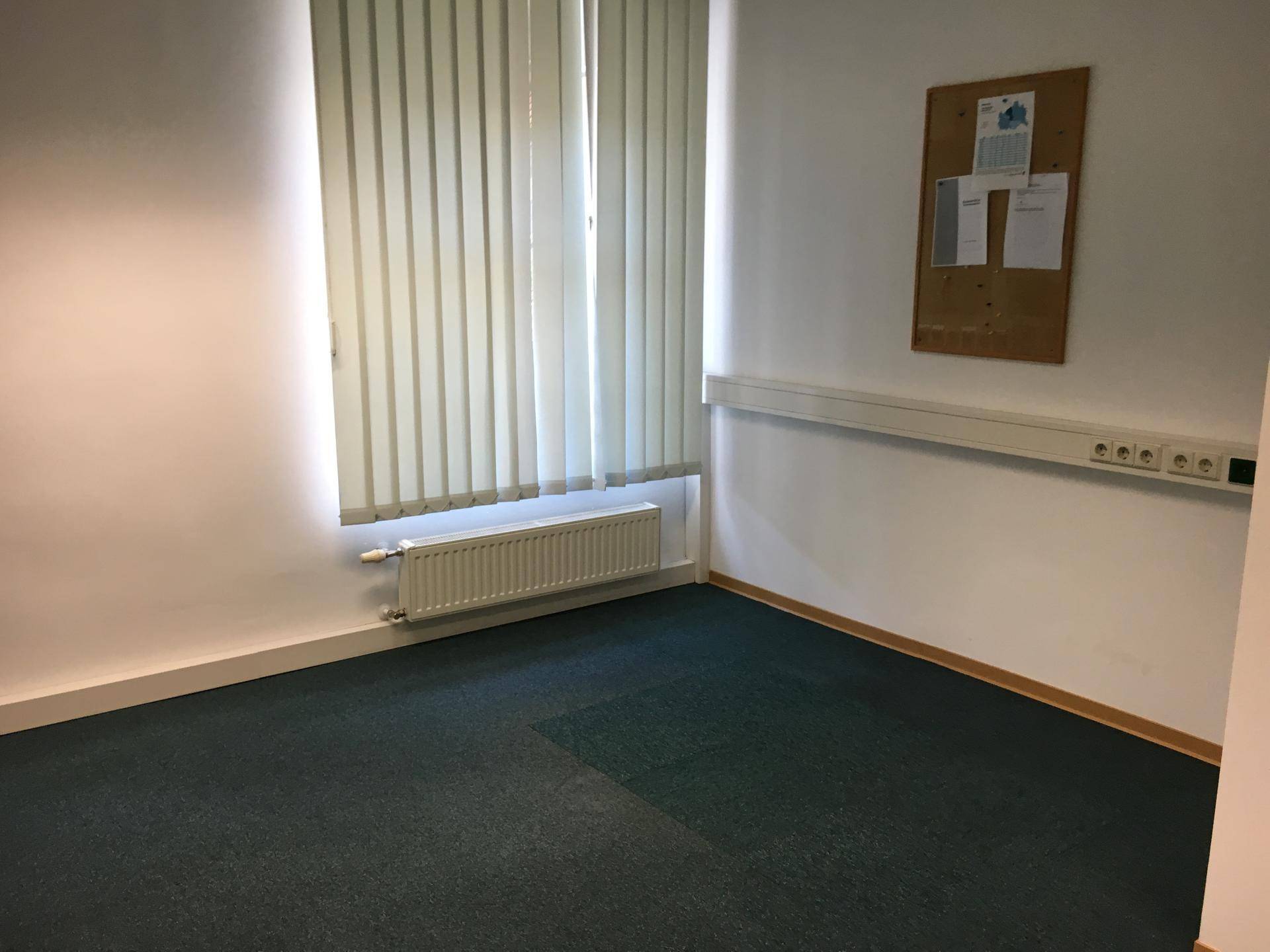 Büroraum mit vertikalen Jalousien, Heizkörper und einer Pinnwand an der Wand.