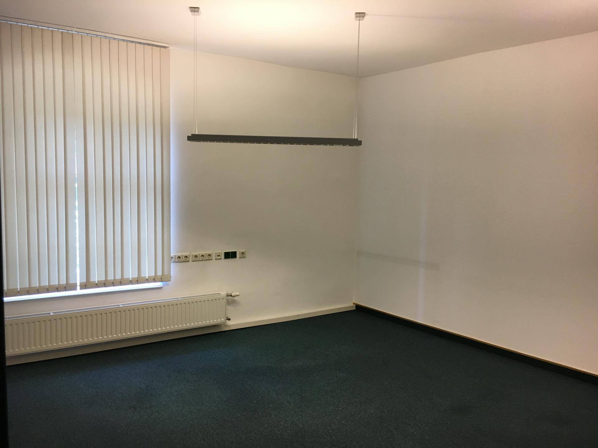 Leerer Büroraum mit vertikalen Jalousien, Heizkörper und moderner Deckenbeleuchtung.