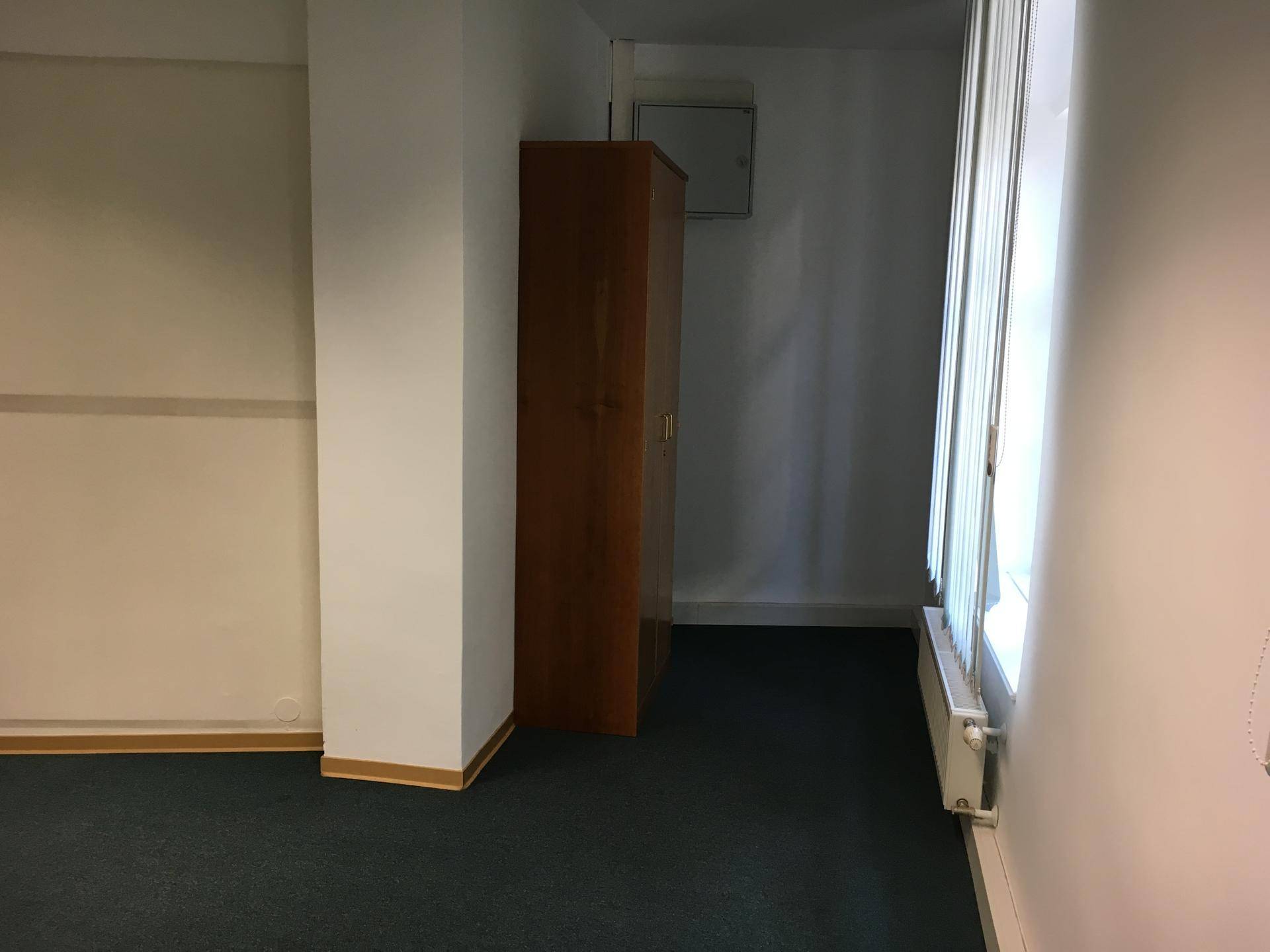 Büroraum mit dunkelgrünem Teppichboden, einem Holzschrank und vertikalen Jalousien.